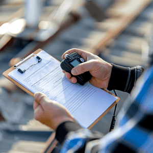 A close-up of a person's hands holding a clipboard with paperwork and an electronic device. The individual, wearing a blue and black jacket, appears to be standing outdoors at an industrial roofing site, suggesting involvement in large-scale roofing projects.