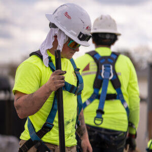 Two construction workers in bright yellow shirts and safety harnesses. One in the foreground wears sunglasses and a white hard hat with a cloth beneath, holding a rod. The other is in the background, focused on their task. Cloudy sky overhead.