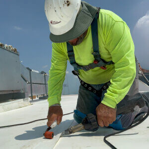 A construction worker in a fluorescent green shirt and hard hat kneels on a rooftop, using tools to work on a  [...]
</p srcset=