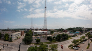 A panoramic view of a cityscape with a large communication tower in the center, surrounded by buildings, streets, and trees. The sky is partly cloudy, and light traffic moves along the roads.