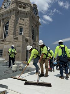 Workers in safety gear and bright vests apply roofing material with brooms on a building rooftop. A clock tower is in the background against a clear  [...]
</p srcset=