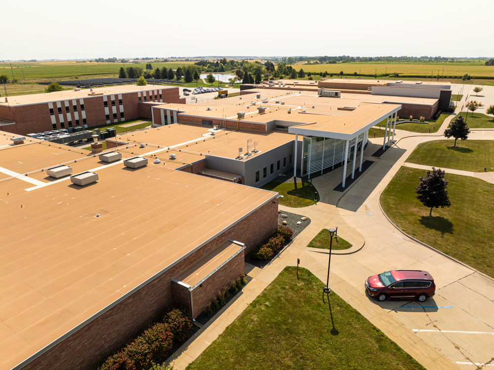 2025-Roof-of-the-Year—Duerson Aerial view of a large, single-story brick building with a tan flat roof, surrounded by green lawns and a few trees. A red car is parked in the driveway near the entrance. The background shows open fields under a clear sky.