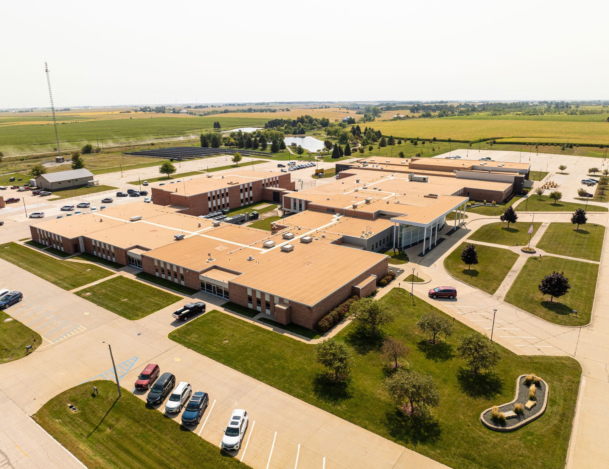 Duerson-Roof-of-the-Year-Grinnell Aerial view of a large school complex surrounded by green lawns and parking lots. The building features multiple connected sections with beige roofs. Cars are parked in designated areas, and fields stretch into the distance under a clear sky.