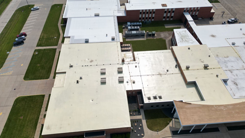 Grinnel-Roof-Before Aerial view of a large building with a white roof, surrounded by grassy areas and parking lots. Several cars are parked in the lot. The structure features multiple sections and varying roof heights.
