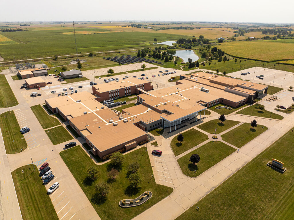 DL2025 Roof of the Year – Duerson-0689 Aerial view of a large, single-story school building with a tan roof, surrounded by parking lots and green fields. Cars are parked in the lots. Nearby, there is a pond and vast farmland stretching into the distance under a clear sky.